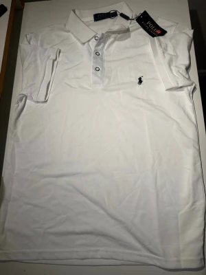 Vit Ralph Lauren pikétröja - Klassisk vit pikétröja från Polo Ralph Lauren med korta ärmar, krage och tre knappar framtill. Tröjan är helt ny och är storlek M.