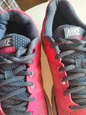 Nike Zoom Winflo rosa sneakers - Snygga Nike Zoom Winflo sneakers i rosa mesh med grå detaljer och vit swoosh. Skorna har grå snörning, vit sula med orange yttersula och ventilerande ovandel. Perfekta för dig som gillar sportig stil och vill ha bekväma skor till vardags.