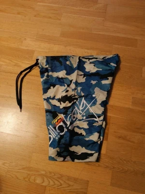 Blå camo shorts från Billabong - Säljer ett par blå camo shorts från Billabong med snörning i midjan och coolt tryck på ena benet. Shortsen har ficka med kardborre på sidan och är perfekta för sommaren. Snygg streetstil och riktigt sköna att bära.
