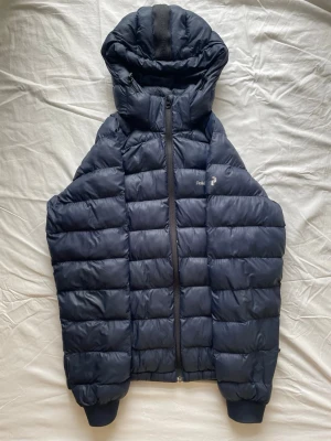 Svart dunjacka från Peak Performance - Snygg svart dunjacka från Peak Performance med huva och dragkedja framtill. Jackan har quiltad design, ribbade muddar vid ärmslut och diskret logga på bröstet. Perfekt för kalla vinterdagar och riktigt varm tack vare dunfyllningen. Och det är exakt som storlek S det är ingen skillnad mellan dom.