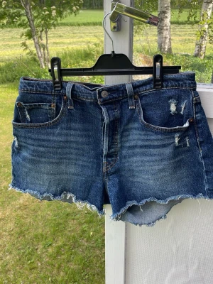 Levi's blå 501 jeansshorts med rå kant - Säljer ett par klassiska blå jeansshorts från Levi's med rå, fransig kant och slitna detaljer framtill. Shortsen har fem fickor, knappgylf och Levi's ikoniska läderpatch bak. Perfekta för sommaren och ger en avslappnad look.