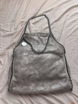 Stella McCartney Falabella handväska - Säljer en ikonisk Stella McCartney Falabella handväska i glittrande silverfärgat veganskt läder. Väskan har kedjedetaljer runt kanterna och axelrem i silverfärgad metall samt en rund logotypbricka. Praktisk dragkedjeficka på baksidan.