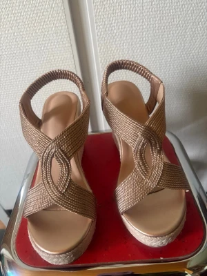 Bronsfärgade kilklackade sandaler, size 37 - Säljer ett par snygga sandaler med kilklack i flätad jute och breda, bronsfärgade band i textil. De har öppen tå och elastiskt band runt hälen för skön passform. Perfekta för dig som vill ha både höjd och komfort i sommarlooken.