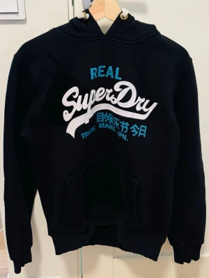Svart Superdry hoodie med tryck - Svart hoodie från Superdry med stor vit och blå logga samt japansk text på bröstet. Klassisk känguruficka framtill, ribbade muddar och justerbar huva med vita snören. Perfekt för en avslappnad streetwear-look. 😁