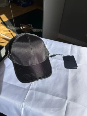 Svart keps i nylon från Prada - Snygg svart keps från Prada i glansigt nylonmaterial med justerbart spänne bak. Klassisk böjd skärm och ikonisk metallogo på sidan. Perfekt accessoar för en trendig och stilren look.