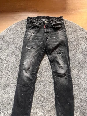 Svarta slitna jeans  - Säljer ett par svarta jeans med slitningar, hål och vita färgstänk för en riktigt edgy look. Jeansen har klassisk femficksmodell, knappgylf och röd logotagg vid fickan. Perfekta för dig som gillar streetstyle och vill sticka ut. Priset går att diskutera