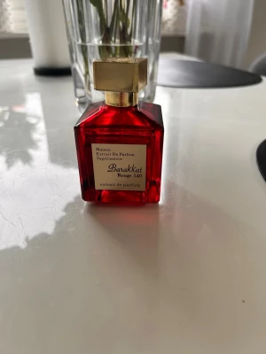 Barakkat Rouge 540 parfym - Lyxig parfym i en fyrkantig röd glasflaska med guldfärgat lock. Flaskan har en elegant etikett framtill och locket är dekorerat med ett ingraverat mönster. Perfekt för dig som gillar exklusiva dofter och snygg design.