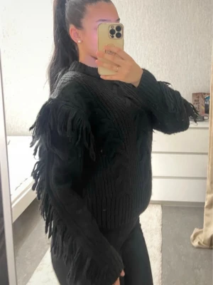 Svart stickad tröja med fransar - Säljer en svart stickad tröja med fransdetaljer längs ärmar och axlar. Tröjan har rund halsringning och är gjord i ett tjockare garn som ger en mysig känsla. Perfekt för dig som gillar statement-plagg och vill sticka ut lite extra.