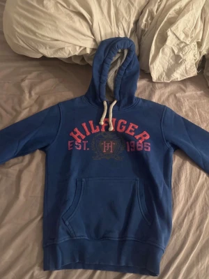 Blå Hilfiger hoodie med tryck - Snygg blå hoodie från Tommy Hilfiger med stort rött tryck på bröstet där det står 'Hilfiger EST. 1985' och ett emblem i mitten. Hoodien har huva med vita snören och en stor magficka. Perfekt för en avslappnad och sportig stil. Jätte bra skick, det står storlek XL men jag skulle säga att det är storlek M.