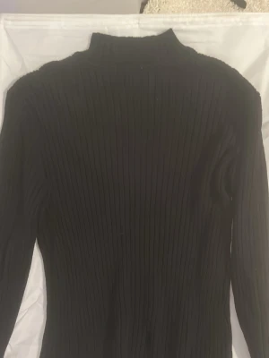 Svart ribbstickad polotröja - Säljer en svart ribbstickad polotröja med lång ärm och hög krage. Tröjan är figurnära och har en klassisk, stilren look som passar till det mesta. Perfekt för lager på lager och enkel att matcha med olika outfits.