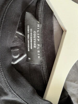 Svart Philipp Plein t-shirt med tryck - Snygg svart t-shirt från Philipp Plein Couture med vita logotryck på ärmarna och ett emblem i nacken. Klassisk rund halsringning och korta ärmar. Tillverkad i mjuk bomull för skön känsla och stilren look.
