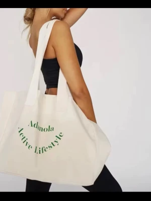 Beige shoppingväska från Adanola - Stor shoppingväska i beige canvas med grönt tryck där det står 'Adanola Active Lifestyle'. Väskan har två långa handtag och rymlig design, perfekt för att bära med sig allt man behöver. Enkel och stilren look som passar till många outfits.