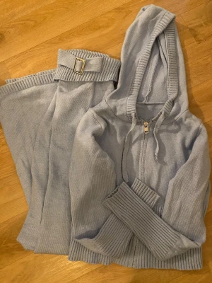 Ljusblått stickat loungewear-set - Mysigt loungewear-set i ljusblått stickat material. Setet består av en zip-hoodie med dragsko och ribbade muddar samt matchande byxor med bred ribbad midja och dekorativt bälte med metallspänne
