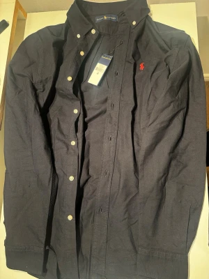 Mörkblå skjorta från Ralph Lauren - Klassisk svart skjorta från Ralph Lauren med röd broderad logga på bröstet. Tröjan är helt ny och är i storlek M. 