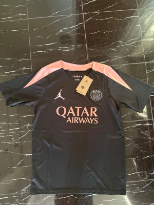 PSG x Jordan svart tränings t-shirt - Snygg svart PSG tränings t-shirt från Jordan med rosa detaljer på axlarna och rosa loggor. Stort Qatar Airways-tryck på bröstet och PSG-emblem. Tillverkad i lätt och snabbtorkande material, perfekt för träning eller match.