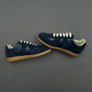 Marinblå sneakers från Maison Margiela - Snygga marinblå sneakers från Maison Margiela med ovandel i mocka och läder, vita snören och klassisk gummisula i brunt. Ikonisk design med diskreta paneler och låg silhuett. Perfekta för dig som gillar stilrena och trendiga skor.