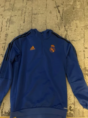 Blå Real Madrid träningströja Adidas - Snygg blå träningströja från Adidas med Real Madrid-logga och Adidas-märke i orange på bröstet. Tröjan har halv dragkedja, svarta ränder på axlarna och är tillverkad i ett tekniskt funktionsmaterial. Perfekt för träning eller uppvärmning.