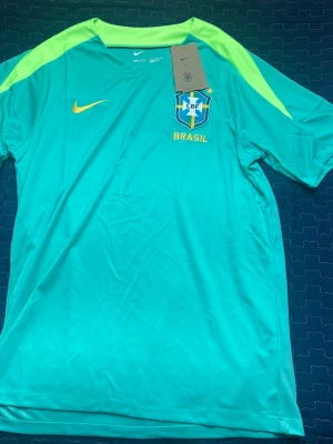 Nike Brasil fotbollströja turkos - Snygg turkos fotbollströja från Nike med Brasil-logga och gula detaljer. Tröjan har korta ärmar, rund hals och är tillverkad i ett lätt, snabbtorkande material som passar perfekt till träning eller match.