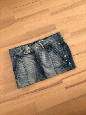 Kort jeanskjol med bälte och knappar - Säljer en snygg kort jeanskjol i blå denim med tillhörande bälte och dekorativa knappar på sidan. Kjolen har fickor fram och bak. Perfekt nu till sommaren!
