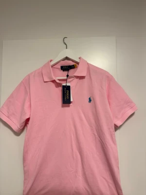 Ljusrosa piké från Polo Ralph Lauren - Snygg rosa pikétröja från Polo Ralph Lauren med klassisk krage och korta ärmar. Tröjan har den ikoniska blå logotypen broderad på bröstet och är tillverkad i mjuk bomull för en bekväm känsla. Perfekt för en avslappnad men stilren look