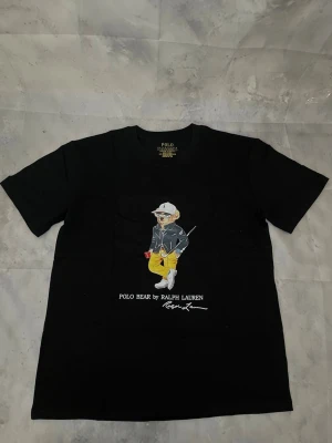 Svart Polo Bear t-shirt från Ralph Lauren - Svart t-shirt från Ralph Lauren med Polo Bear-tryck framtill. Klassisk passform med rund hals och korta ärmar. Tillverkad i mjuk bomull och har vit text under björnmotivet. Perfekt för en avslappnad och stilren look.