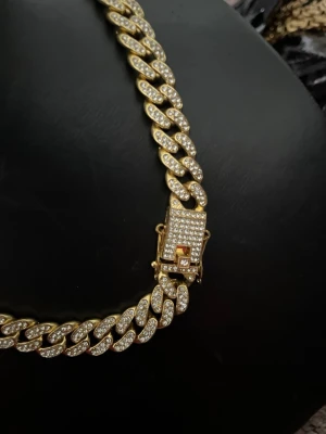Guldfärgat cuban chain halsband - Snyggt guldfärgat cuban chain halsband med glittrande stenar på varje länk. Halsbandet har en rejäl låsdel som också är dekorerad med stenar för extra bling. Perfekt för dig som vill sticka ut och addera lite extra shine till din outfit.