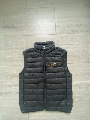  EA7 väst - Snygg svart väst från Emporio Armani EA7 med guldfärgad logga på bröstet.Perfekt för en sportig vibe till din outfit.