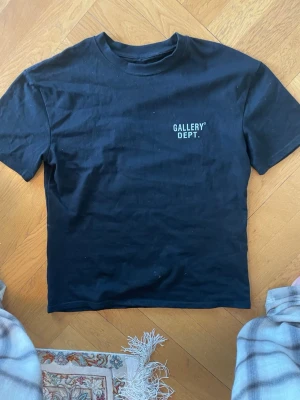 Svart t-shirt från Gallery Dept. - Svart t-shirt från Gallery Dept. med tryck på bröstet och stor logga på ryggen. Klassisk passform med rund hals och korta ärmar. Perfekt för en avslappnad och trendig stil.