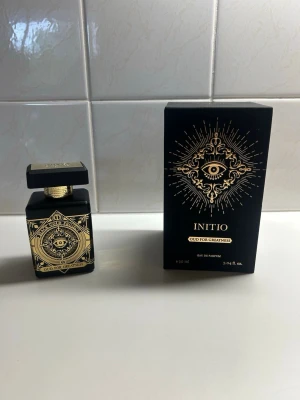 Initio Oud For Greatness parfym - Initio Oud For Greatness är en exklusiv parfym i en svart, fyrkantig flaska med detaljer i guld och ett mystiskt öga på etiketten. Flaskan har en lyxig känsla med geometriska mönster och matchande svart och guld förpackning. Eau de Parfum, 90 ml.