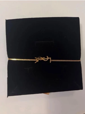 Guldigt YSL armband - Snyggt och stilrent armband i guldton från Yves Saint Laurent. Tunt kedjearmband med YSL-logga i mitten som detalj. Perfekt accessoar för dig som gillar minimalistisk och exklusiv design. Passar till många olika stilar och är lätt att matcha.
