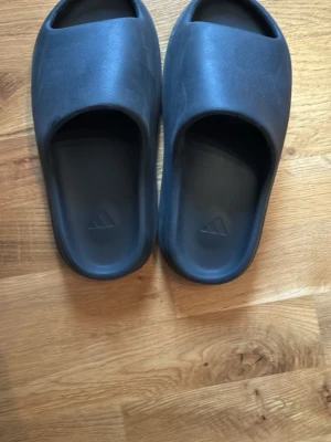 Slides (tofflor) - Säljer ett par mörkgråa yeezy slides med stilren design och öppen tå. Tillverkade i ett mjukt och bekvämt syntetmaterial med Adidas-logga på fotbädden. Perfekta för chill dagar hemma eller till och från träningen. Priset är inte hugget i sten och säljes pga att de är för små