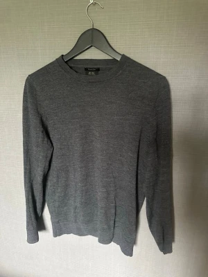 Mörkgrå stickad tröja från Massimo Dutti - Snygg mörkgrå stickad tröja från Massimo Dutti i mjuk ullblandning. Tröjan har rund halsringning, långa ärmar och en klassisk, rak passform. Perfekt att matcha med jeans eller chinos för en clean look.