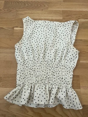 Vit prickig topp från Gina Tricot - Supersöt vit topp från Gina Tricot med svarta prickar. Toppen är ärmlös, har smock i midjan och en volangkant nedtill som ger en extra feminin touch. Perfekt för dig som gillar en lekfull och trendig stil.