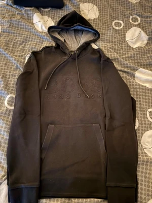 Svart hoodie från Hugo Boss - Snygg svart hoodie från Hugo Boss med diskret broderad logga på bröstet. Hoodien har en grå insida i huvan, dragsko och en stor magficka. Perfekt för en avslappnad och stilren look. Materialet känns mjukt och bekvämt.