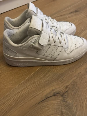 Vita Adidas sneakers med kardborre - Snygga helvita sneakers från Adidas med klassiska tre ränder på sidan, perforerad tå och kardborreband över snörningen. Skorna är låga och har en stilren design i läder med Adidas-logga på sidan och plös. Perfekta för en clean och sportig look.