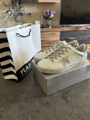 Golden Goose Skor - Snygga vita sneakers från Golden Goose med grå stjärndetalj på sidan och svart hälparti med logga. Skorna har grå skosnören och en slitstark sula i gummi. Klassisk låg modell i läder som passar perfekt till streetwear och casual outfits.