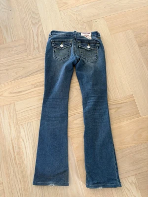Blå bootcut jeans från True Religion - Säljer ett par blå bootcut jeans från True Religion med klassiska kontrastsömmar och snygga fickdetaljer bak. Jeansen har låg midja, silverfärgade knappar och är tillverkade i stretchigt denimtyg för skön passform. Lite slitage nertill på benen. Se bild 