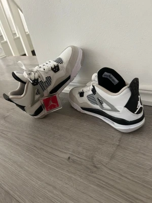 Air Jordan 4 Retro White/Black - Snygga Air Jordan 4 Retro sneakers i vitt och svart med meshdetaljer och klassisk Jumpman-logga på hälen. Skorna har vita skosnören, svarta detaljer och extra snören medföljer. Tillverkade i läder och mesh för en sportig look.