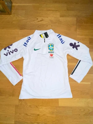 Vit Brasil Nike träningströja - Vit långärmad träningströja från Nike med Brasiliens landslagslogga och sponsorer som Itaú och Guaraná Antarctica. Halvlång dragkedja framtill och tryck i blått, grönt och lila på ärmar och bröst. Perfekt för fotbollsträning eller sportig stil.