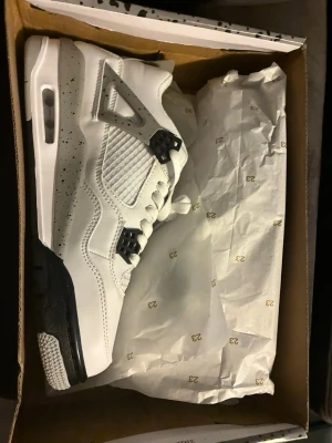 Nike Air Jordan 4 White Cement - Grymma Nike Air Jordan 4 White Cement sneakers i vitt läder med grå och svarta detaljer. Klassisk siluett med meshpaneler, synlig Air-sula och speckle-mönster på mellansulan och hälen. Perfekta för dig som vill sticka ut med en ikonisk och tidlös sneaker.