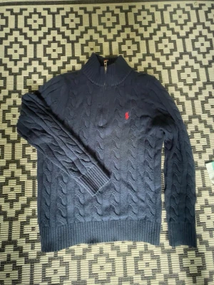 Mörkblå kabelstickad tröja Polo Ralph Lauren - Mörkblå kabelstickad tröja från Polo Ralph Lauren med halv dragkedja och hög krage. Tröjan har ett rött broderat logomärke på bröstet och ribbstickade muddar. Perfekt för en stilren och klassisk look.