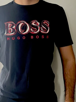 Svart Hugo Boss t-shirt med tryck - Snygg mörkblå t-shirt från Hugo Boss med stort grafiskt BOSS-tryck i vitt och rött på bröstet. Klassisk passform och rund halsringning. Tillverkad i mjuk bomull som känns skön mot huden. Perfekt för dig som vill ha en stilren men ändå iögonfallande look. Storleken är L men den har endast passform för M, den är ÄKTA.