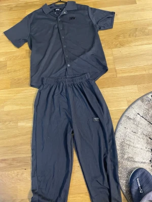Mörkgrått set med skjorta och byxa - Säljer ett mörkgrått set från SikSilk med kortärmad skjorta med knappar och matchande byxa med resår i midjan. Båda delarna har en liten svart logotyp-patch. Materialet är mjukt och lätt, perfekt för en avslappnad look.