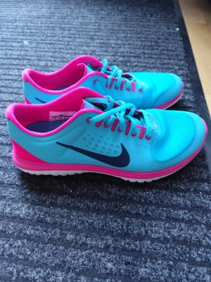 Nike turkosa och rosa sneakers - Nike sneakers i en fräsch turkos färg med rosa detaljer och innersula. Skorna har en låg profil, snörning och vit sula. Swoosh-loggan i mörkblått på sidan. Ovandelen är i mesh och syntetmaterial för bra andningsförmåga.