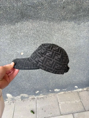Svart keps med Fendi-logga - Säljer en svart keps från Fendi med det klassiska FF-monogrammet över hela kepsen. Kepsen har böjd skärm, justerbar rem bak och är tillverkad i ett tygmaterial. Perfekt för dig som vill ha en stilren och lyxig accessoar.
