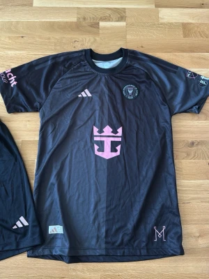 Svart Inter Miami matchtröja och shorts Adidas - Säljer ett svart Inter Miami set från Adidas med rosa detaljer och klubbmärke på bröstet. Tryck Messi på ryggen. Tröjan har rund hals, korta ärmar och flera loggor i rosa och vitt. Ny och ej använd💞