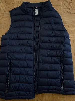 Mörkblå dunväst från U.S. Polo Assn. - Snygg mörkblå dunväst från U.S. Polo Assn. med quiltad design och diskret logga på bröstet. Västen har hög krage, dragkedja framtill och två fickor med dragkedja. Perfekt för lager-på-lager och ger en sportig vibe till din outfit.