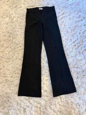 Svarta utsvängda kostymbyxor - Snygga svarta kostymbyxor med utsvängda ben och hällor för skärp. Klassisk midja och stilren design som passar perfekt till en trendig look. Materialet ser ut att vara en mjuk och följsam mix, perfekt för dig som gillar stilrena och bekväma plagg. Som nya!