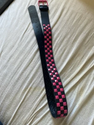 Svart bälte med rosa nitar - Säljer ett svart bälte med rosa fyrkantiga nitar i rutmönster. Bältet har en rosa metallspänne och är perfekt för att ge din outfit en alternativ vibe. Passar dig som gillar att sticka ut och vill ha en cool accessoar.