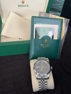 RoIex Datejust silver herrklocka - Snygg RoIex Datejust herrklocka i silver med klassisk jubilee-länk och räfflad bezel. Urtavlan är grå med tydliga index och datumfönster. Klockan kommer med originalbox, certifikat och tillbehör. Perfekt för dig som vill ha en tidlös och lyxig accessoar.